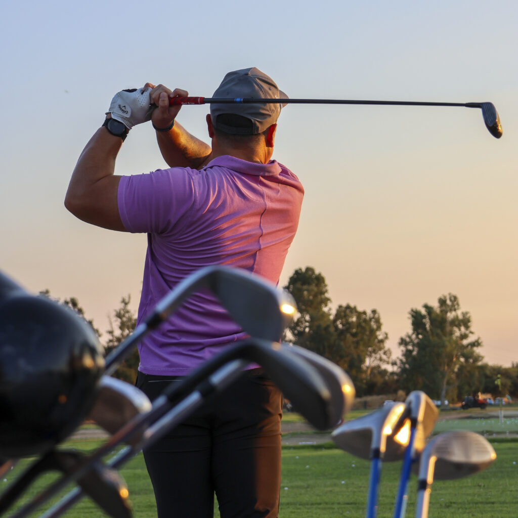 Golfer finishing a swing at sunset on a driving range, with golf clubs in the foreground and practice targets visible in the distance.