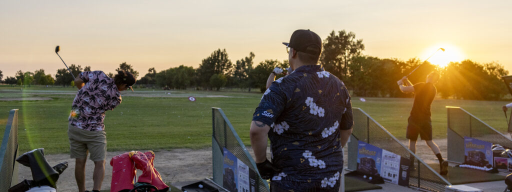 People enjoying an evening at a golf driving range during sunset, with golfers taking swings in adjacent bays while others relax and socialize. The warm golden light, golf clubs, and open range create a casual after-work or weekend entertainment atmosphere.