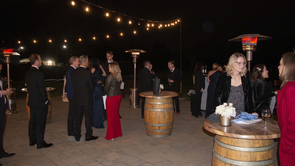 Guests mingling on an outdoor wedding reception patio at night with string lights, cocktail tables, wine barrel tables, and patio heaters.