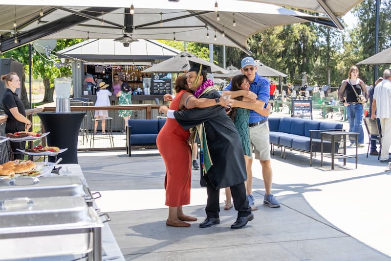Graduate in cap and gown hugs family members at an outdoor patio beside a golf driving range, with food service and seating areas in the background.