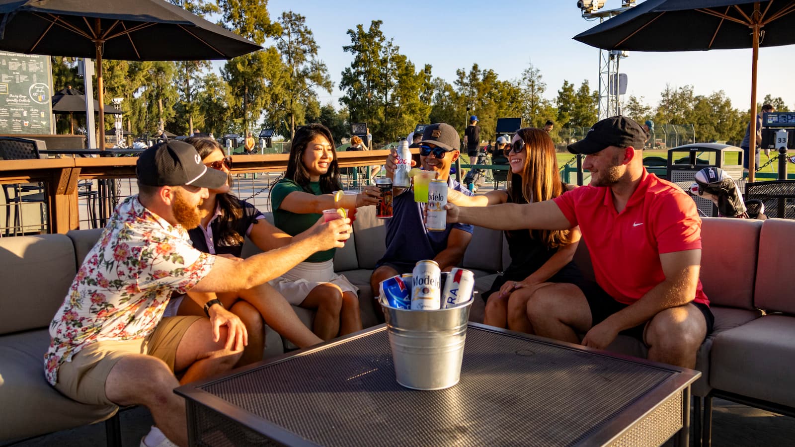 Group of friends toasting with drinks while relaxing in a reserved bay at The Hangout driving range at Haggin Oaks Golf Complex in Sacramento.