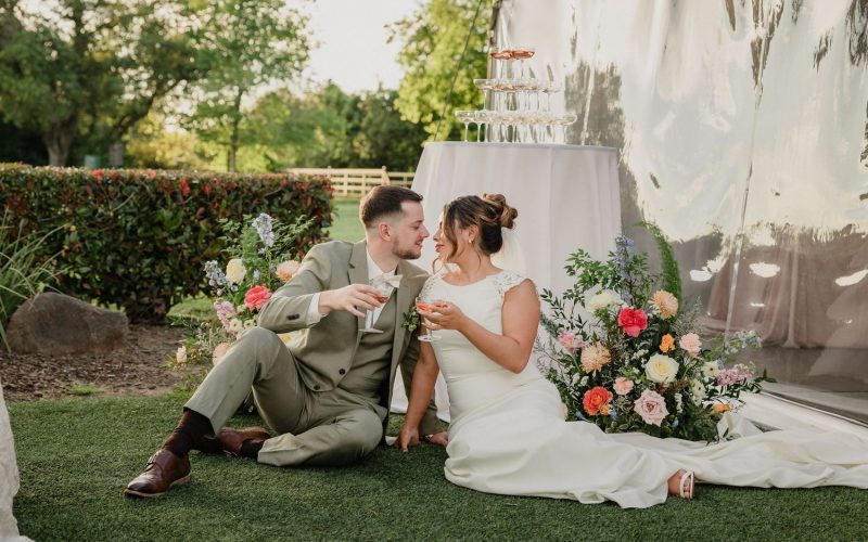 Bride and groom sharing a toast beside floral arrangements outside a reception tent at The Pavilion at Haggin Oaks in Sacramento.